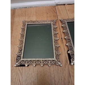 May include: Two ornate, rectangular picture frames with gold-colored filigree detailing and faux pearl accents. The frames have a dark green interior and sit on a wooden surface. The frames are likely vintage or antique.