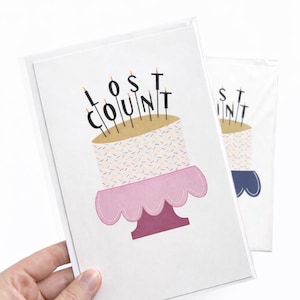 May include: Two birthday cards in plastic sleeves. The cards feature a cake design with the words "LOST COUNT" in black letters above the cake. The cake has a pink cake stand and is decorated with sprinkles and candles.