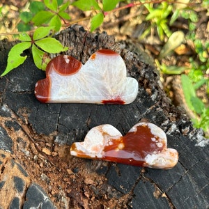 May include: Two agate cloud shaped stones with white and red patterns. The stones are on a brown wooden surface.