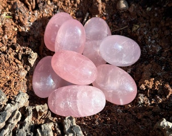 May include: A cluster of smooth, oval-shaped rose quartz stones. The stones are a soft pink color and appear translucent. They are nestled in a dark, textured wooden surface, creating a natural and earthy aesthetic.