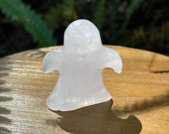 May include: A white selenite ghost figurine with a carved face. The ghost is standing on a wooden surface.