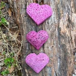 May include: Three pink heart-shaped decorations made of a porous material, arranged vertically on a tree trunk.