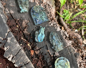 May include: Five small carvings of various figures, including an angel, a flower, a bird, a person, and a Buddha, are displayed on a piece of weathered wood. The carvings are made of iridescent labradorite stone, which shimmers with shades of green, blue, and gold.