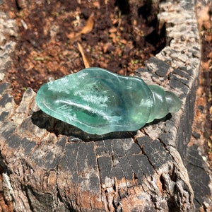 May include: A green fluorite crystal carved into the shape of a seashell. The crystal is translucent and has white veins running through it. The crystal is sitting on a piece of weathered wood.