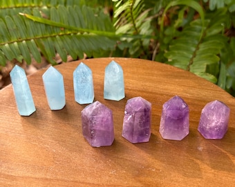 May include: A collection of polished crystal points in shades of light blue and purple. The crystals are arranged on a wooden surface, with green foliage in the background. The crystals are likely used for decorative or spiritual purposes.