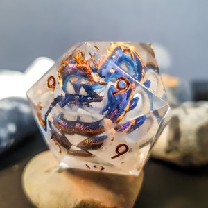 以下が含まれることがあります： 青と金の龍のデザインが施された、透明で虹色の樹脂製のd20ダイス。ダイスには、3つの面に9の数字が刻まれています。