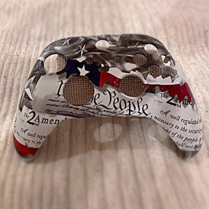 May include: A white and gray video game controller skin with a red, white, and blue American flag design. The skin features the text "We the People" and "The 2nd Amendment".