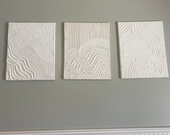 May include: Three white textured canvas paintings with abstract wavy lines hanging on a light green wall above a white wainscoting.