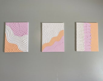 May include: Three abstract paintings with a textured, layered design. The paintings feature a combination of white, pink, and peach colors.
