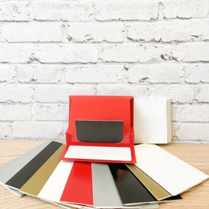 May include: A stack of red, white, gold, black, and silver cardstock paper. The top cardstock is red and has a black rectangle in the center. The cardstock is folded in half and has a white rectangle in the center.