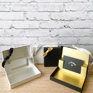 May include: Three gift boxes with black, white, and gold accents. The box on the right has a gold interior and a black gift card with the text "Callaway" in white.
