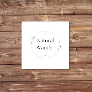 May include: A square white card with the words "Natural Wander" in a circular design, featuring delicate leaf and butterfly illustrations. The card is set against a rustic, brown wooden background.