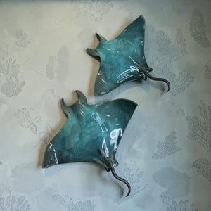 Op de afbeelding: Twee keramische wandsculpturen in de vorm van mantaroggen, voorzien van een glanzend blauw en groenblauw glazuur met een subtiel bubbelachtig textuurpatroon. Elke rog heeft brede, platte vinnen en een lange, gebogen staart.