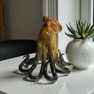 May include: A glass octopus figurine featuring a gradient color scheme that transitions from a warm amber and brown tone at the head to a deep, translucent charcoal gray at the tips of its eight curled tentacles, showcasing a smooth finish.