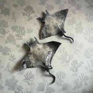 Op de afbeelding: Twee wandsculpturen in de vorm van een rog met een glanzende afwerking. Elk stuk heeft een verloop van zwarte, grijze en witte tinten met een opvallend bubbelachtig patroon over de vleugels en een gebogen, slanke staart aan het lichaam.