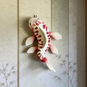 May include: A wall-mounted koi fish sculpture featuring a glossy white body with vibrant red, organic-shaped markings along the back. The fish has detailed fins, a defined tail, and small dark eyes, presented in a swimming position with a smooth finish.