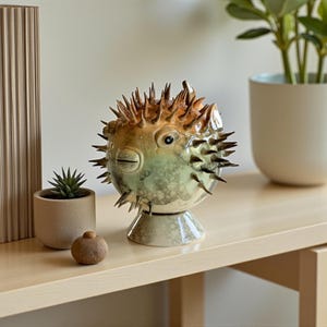 May include: A ceramic pufferfish sculpture with a spiky exterior, featuring shades of green, yellow, and brown. The decorative piece has a detailed face and sits on a pedestal base. The sculpture is displayed on a light-colored wooden surface.