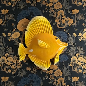 Puede incluir: Una vibrante escultura de pez amarillo con una prominente aleta dorsal y aleta caudal. El pez tiene un acabado brillante y está colocado sobre un fondo oscuro con un patrón floral. Es un objeto decorativo.