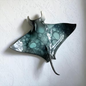 May include: A ceramic wall sculpture shaped like a manta ray, featuring a glossy finish with a gradient of deep charcoal to teal. The surface displays a distinct bubble-like pattern in varying shades of blue and green, with a long, slender tail.