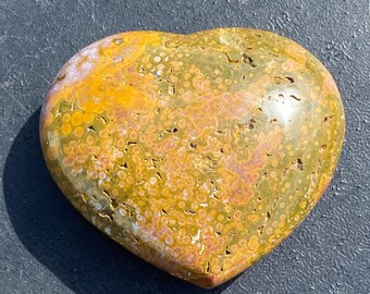 May include: A heart-shaped stone with a speckled pattern in shades of yellow, green, and brown. The polished surface reflects light, highlighting the natural variations in color and texture. The stone is smooth and rounded, with a slight sheen.