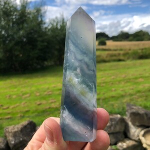 Könnte beinhalten: Eine blaue, grüne und weiße Fluorit-Kristallspitze. Der Kristall ist klar und hat eine glatte, polierte Oberfläche. Es ist ein Naturstein und hat ein einzigartiges, wirbelndes Muster.