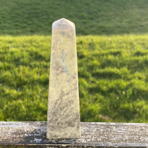 May include: A gray, green, and white obelisk shaped crystal with a pointed top. The crystal has a mottled, cloudy appearance.