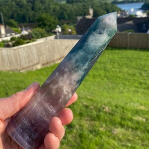 Könnte beinhalten: Eine grüne, violette und weiße Fluorit-Kristallspitze. Der Kristall ist glatt und poliert, mit einer spitzen Spitze. Es ist ein natürlicher Kristall, und die Farben sind lebendig und einzigartig.