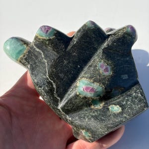 May include: A dark green and black carved stone sculpture with ruby and emerald accents. The sculpture has a geometric shape with multiple peaks and valleys. The stone has a polished finish and is held in a hand.