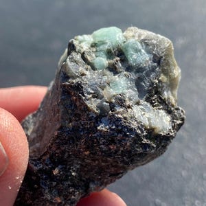 以下が含まれることがあります： 鮮やかな緑色の原石エメラルド。暗く、質感のある岩の母岩に埋め込まれています。エメラルドの結晶が自然な形で確認できます。手で持っており、そのサイズと詳細が際立っています。