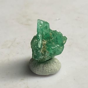 May include: A cluster of green emerald crystals on a white display stand.