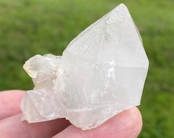 May include: A clear quartz crystal cluster with a large, pointed main crystal and smaller crystals attached. The quartz is translucent with a slightly milky appearance, held in a hand against a blurred green background.