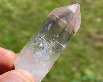 May include: A clear quartz crystal point with a light pink hue and internal inclusions. The crystal is pointed at one end and has a flat base.