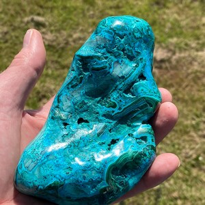 May include: A large, polished stone with a vibrant mix of teal and green hues. The stone's surface has intricate patterns and textures, creating a unique and eye-catching appearance. The stone is held in a hand, showcasing its size and shape.