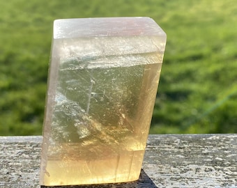 May include: A large, clear, rectangular crystal with a yellow and pink hue. The crystal has a smooth, polished surface and is sitting on a wooden surface.