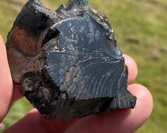 May include: A black obsidian rock with a striated surface. The rock has a rough, uneven texture and a dark, glossy finish.