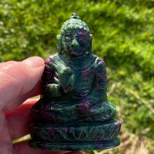 May include: A carved Buddha statue in a seated position, featuring a lotus base. The statue is a mix of green and ruby red colors. The Buddha's hands are in a prayer position, and the statue is set against a blurred green background.