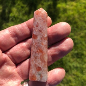 May include: A sunstone point, also known as a sunstone wand, is a crystal with a reddish-orange colour and a translucent appearance. The crystal has a pointed tip and a flat base. It is held in a hand for size reference.