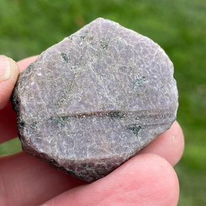 Può includere: Una gemma di rubino naturale, tagliata grezza. La pietra ha una tonalità rosso-violacea smorzata con una texture superficiale visibile e piccole inclusioni verdi. Il rubino è tenuto in mano su uno sfondo verde sfocato.