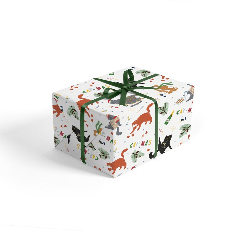 Puede incluir: Papel de regalo blanco con un patr&oacute;n festivo de gatos, &aacute;rboles de Navidad y el texto "Cat-mas". El papel de regalo est&aacute; atado con una cinta verde.