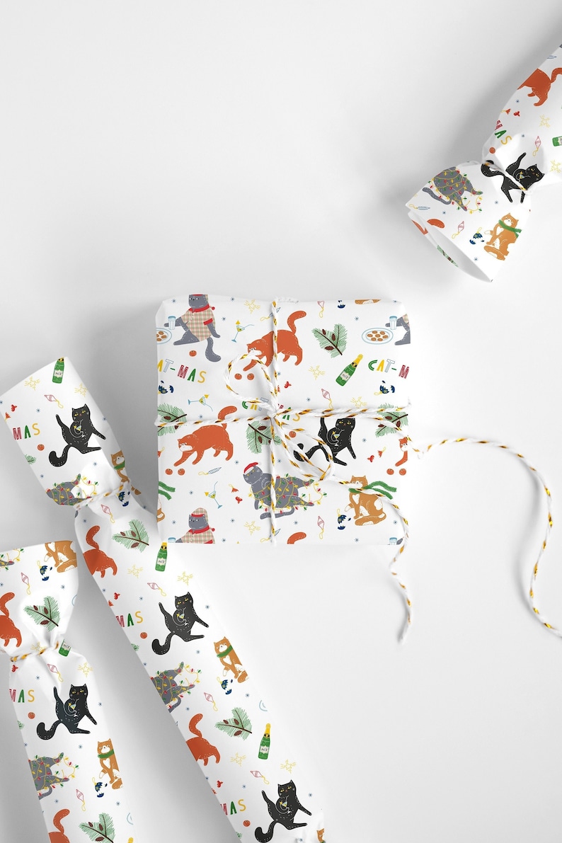 Puede incluir: Papel de regalo blanco con un patr&oacute;n repetido de gatos con gorros de Pap&aacute; Noel, sosteniendo copas de champ&aacute;n y con luces de Navidad. El texto "Cat-mas" se repite en todo el patr&oacute;n.