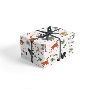 Puede incluir: Papel de regalo blanco con un patr&oacute;n repetitivo de gatos, adornos y el texto "Cat-mas". El papel de regalo est&aacute; atado con una cinta negra.