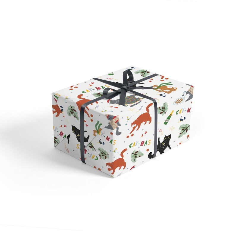 Puede incluir: Papel de regalo blanco con un patr&oacute;n repetitivo de gatos, adornos y el texto "Cat-mas". El papel de regalo est&aacute; atado con una cinta negra.