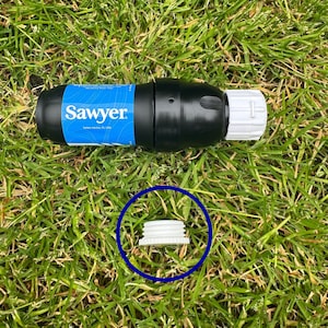 Op de afbeelding: Een zwarte waterfilter met een witte dop en een blauw label met de tekst "Sawyer" en "Safety Harbor, FL USA". De filter ligt op groen gras. Een witte schroefdop is blauw omcirkeld.
