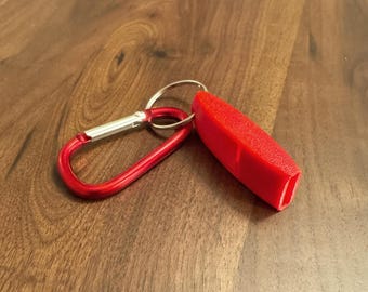 Op de afbeelding: Een rode sleutelhanger met een bijpassende rode karabijnhaak en een klein, rechthoekig, getextureerd rood object. De sleutelhanger ligt op een houten oppervlak.