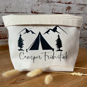 Könnte beinhalten: Eine weiße Stofftasche mit einer schwarzen Silhouette eines Zeltes, Bergen und Bäumen. Der Text "Camper Frühstück" ist unter dem Bild gedruckt.