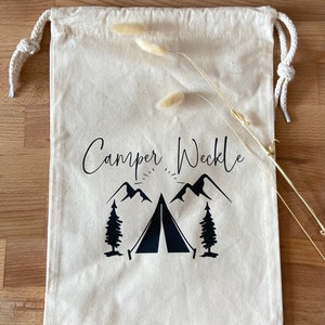 Könnte beinhalten: Eine weiße Stofftasche mit Kordelzug, mit schwarzem Text, der "Camper Weekle" lautet, und einer schwarzen Illustration eines Zeltes, Bergen und Bäumen.