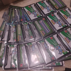 Puede incluir: Una pila de sobres de refuerzo de cartas coleccionables de Pokémon sellados con un fondo verde y negro. Los sobres presentan una imagen holográfica de un personaje Pokémon.