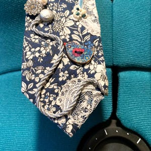 May include: A handmade necktie with a blue floral design, embellished with lace, decorative buttons, and a bird-shaped motif. The tie includes a pale blue ribbon and silver rope detailing.