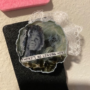 May include: A handcrafted resin brooch with a grayscale portrait. The brooch has a pale blue edge, embellished with white lace. The text "THERE'S NO LEAVING HERE" is printed on a white strip. The brooch is set on a black surface.