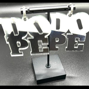 May include: A pair of silver mirror acrylic earrings with the word "DO DO PEPE" written on them. The earrings are hanging from a black stand.